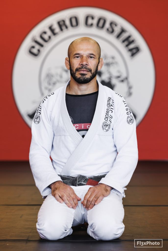 Retrato do Mestre Cícero Costha ajoelhado de kimono, diante do emblema da equipe